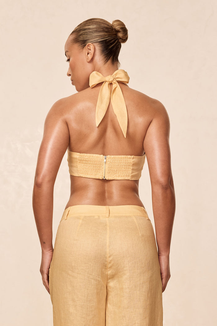 Rear view of Montserrat Top in raffia linen on model, showing halter bow tie and smocked back with center zipper against beige background