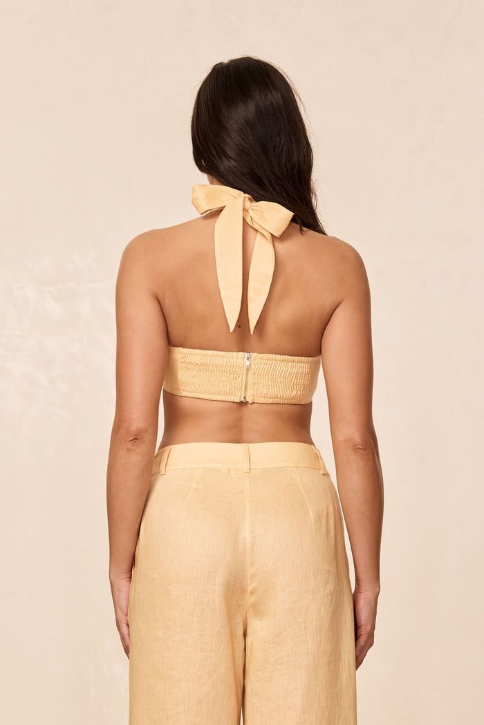 Back view of Montserrat Top in raffia linen showing smocked band with zipper and halter neck bow on beige background