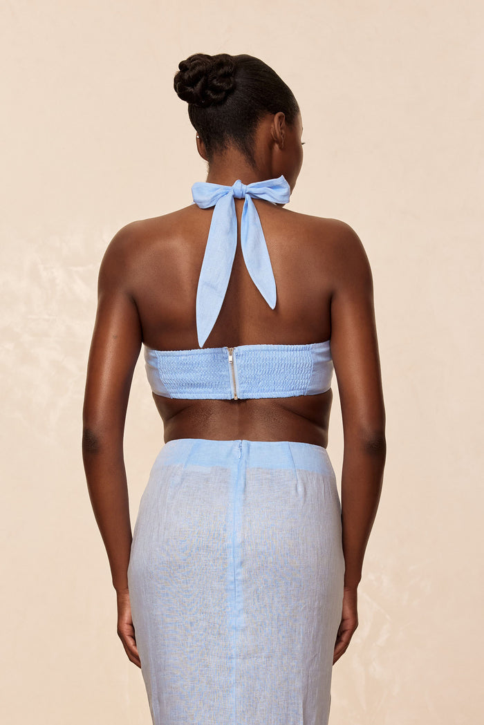 Back view of Montserrat Top in Sky Linen showing halter tie, smocked back band, and cropped fit on model against beige background
