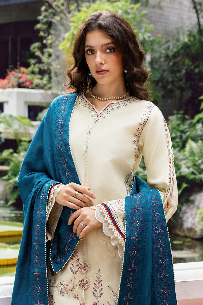 Woman wearing a cream embroidered outfit with a blue shawl outdoors.
