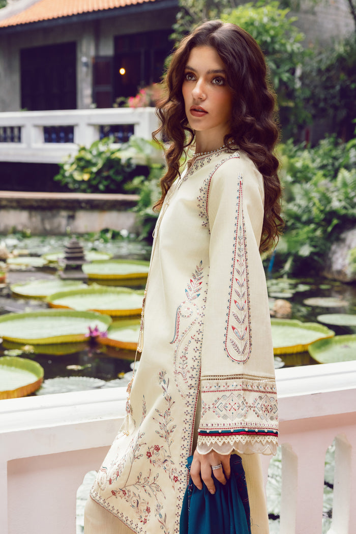 Woman in a cream embroidered outfit standing by a pond with lily pads.
