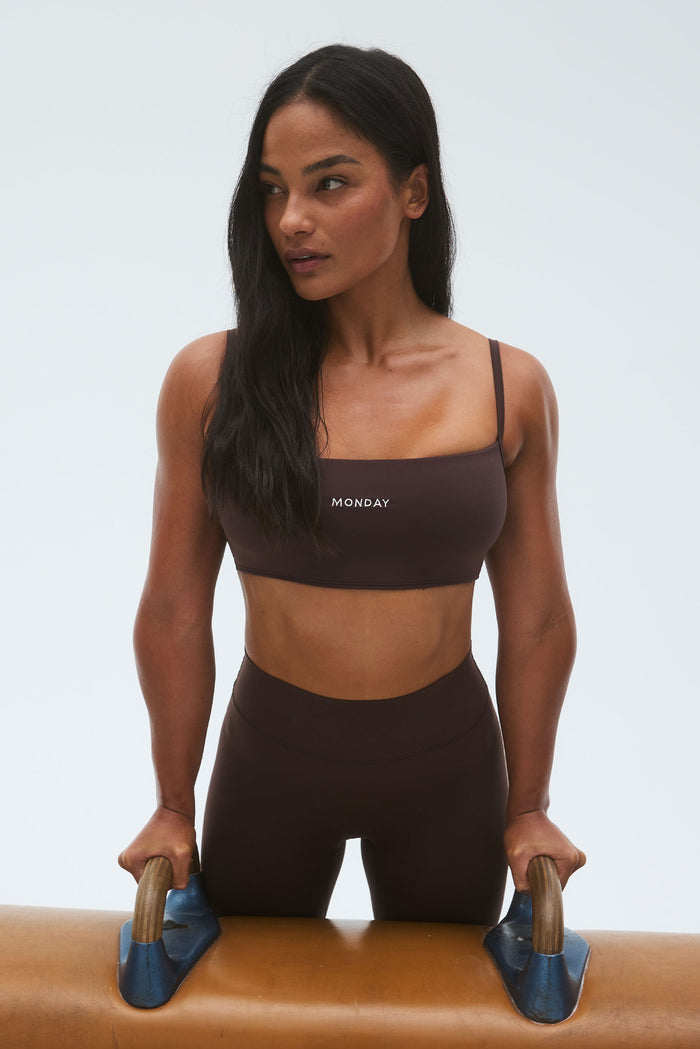 Three-quarter front view of Olympic Bra in Espresso showcasing thin adjustable straps and centered MONDAY logo, model holding pommel horse handles