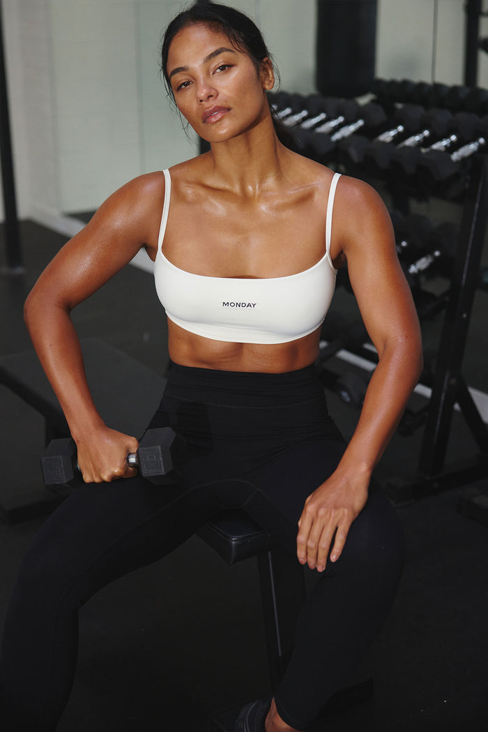 Front view of Olympic Bra in ivory showing thin straps, low scoop neckline, and center MONDAY logo with model seated in gym