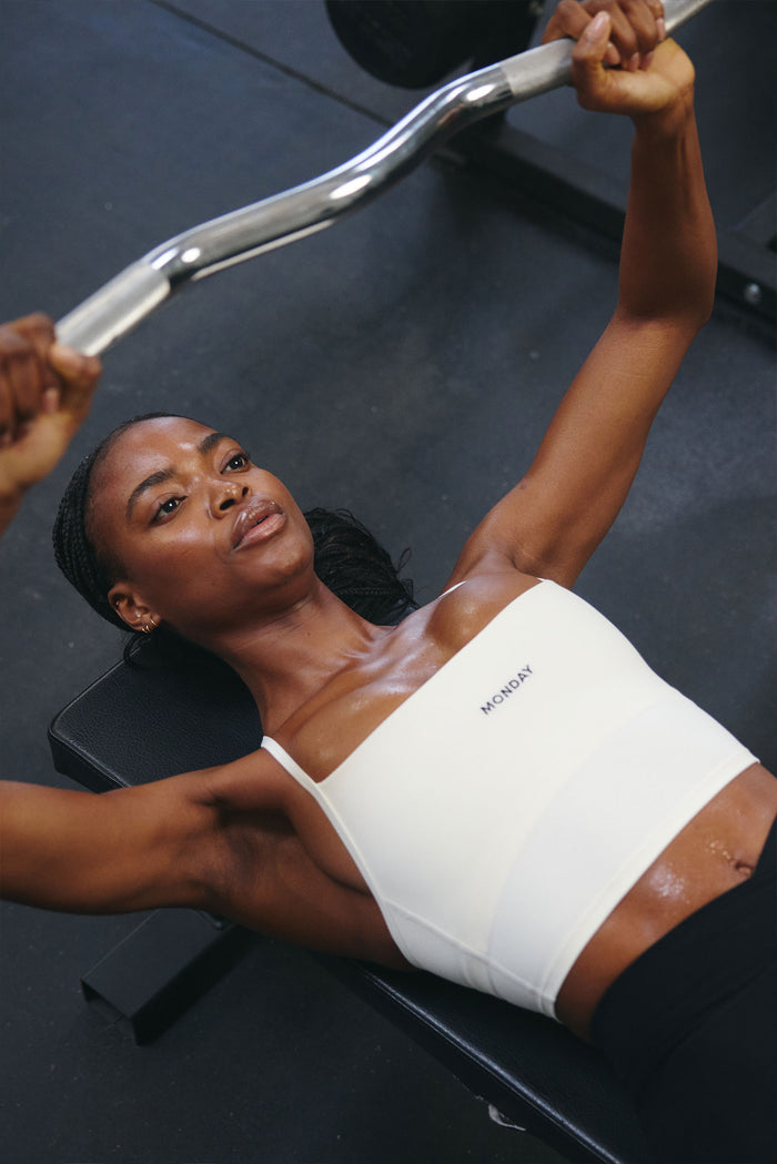 Model bench pressing wearing an ivory Olympic Tank, cropped fit with scoop neckline and centered MONDAY logo in a gym