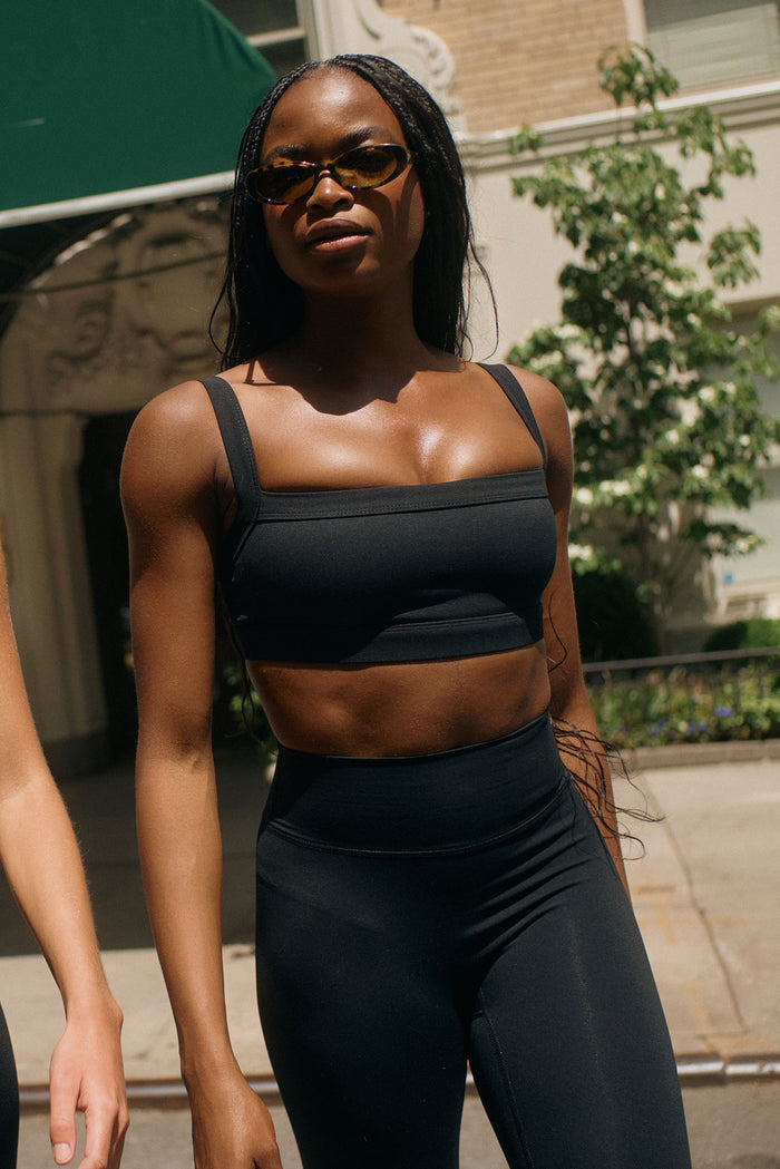 Front view of Outpost Bra in black showcasing straight neckline and wide adjustable straps with sunlit city street background