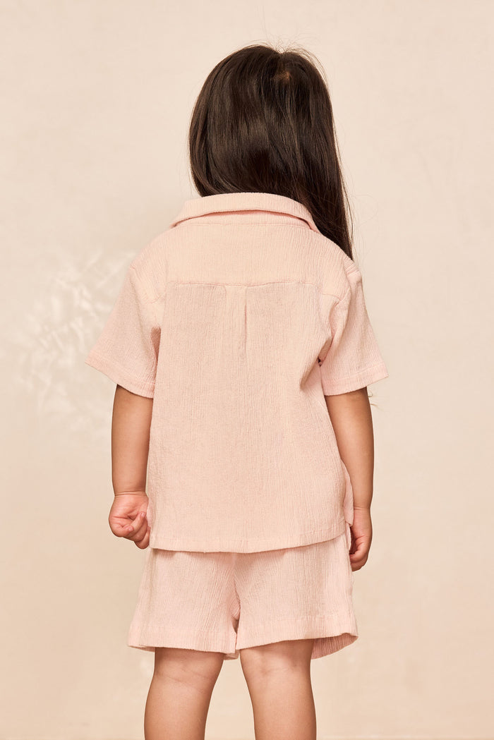 Back view of Pacific Beach Shirt Kids in powder pink showing crinkled texture and short sleeves on model against beige backdrop