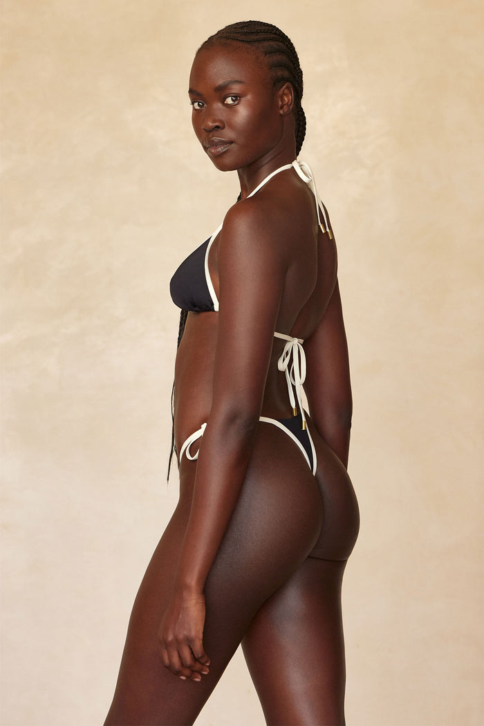 Profile view of Palma Thong in Black/Ivory showcasing high-cut thong back and tie-side straps with beige backdrop