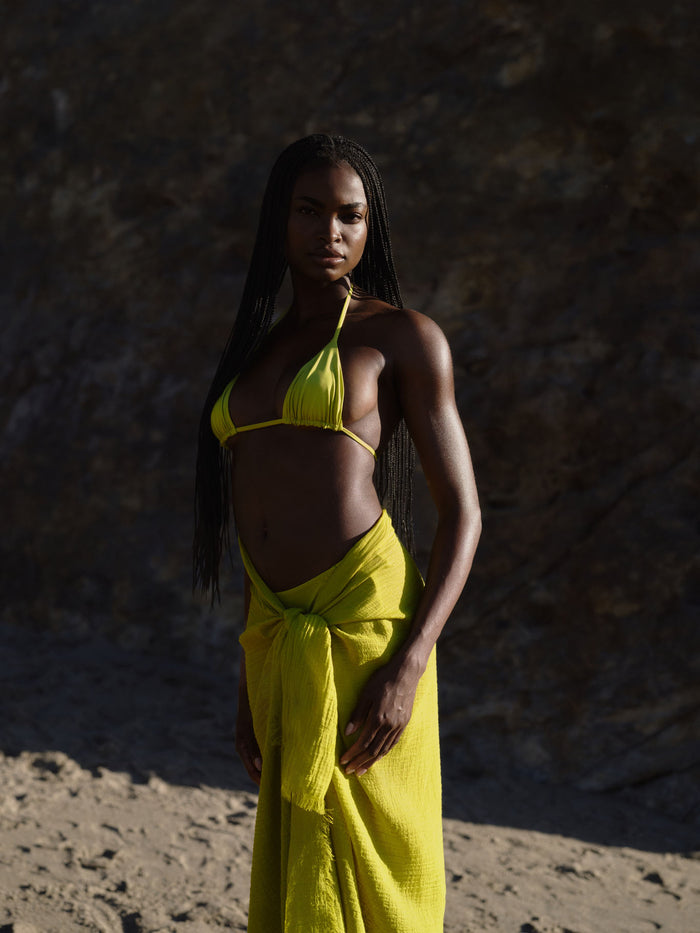 Three-quarter front view of Palma Top in Apple showcasing triangle cups and halter neck ties with rocky beach background