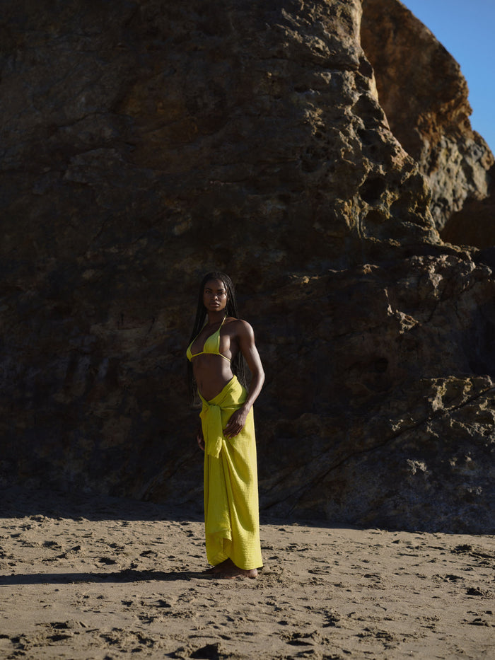 Model wearing Apple Palma triangle bikini top with matching sarong standing on a sandy beach in front of rocky cliffs