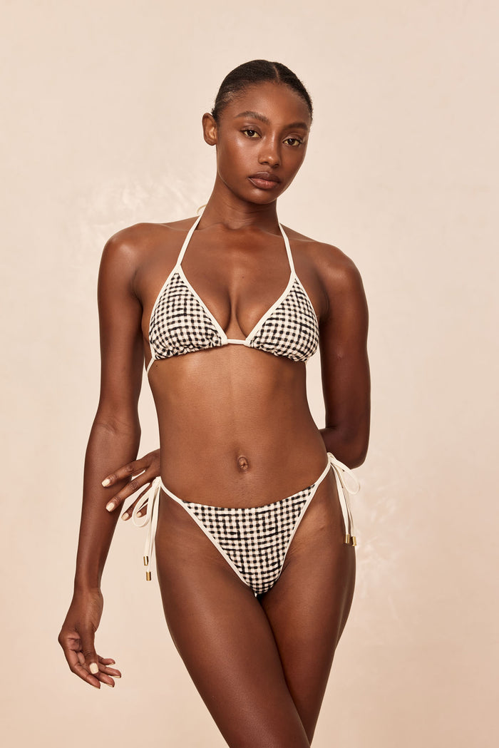 Woman wearing black-and-white gingham tie-side bikini with triangle top, standing against a neutral beige background.
