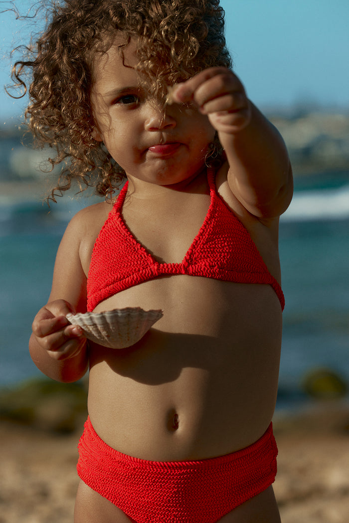 Close-up of Palma Top and Seychelles Bottom in Chili Pepper Crinkle on toddler, red textured knit set holding scallop shell on beach