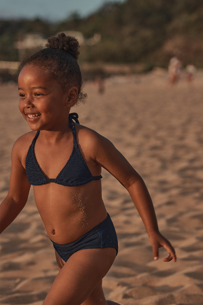 Three-quarter view of Palma Top and Seychelles Bottom set in Midnight Crinkle showing crinkle texture, halter tie top, and mid-rise bottoms on a child running on a sandy beach