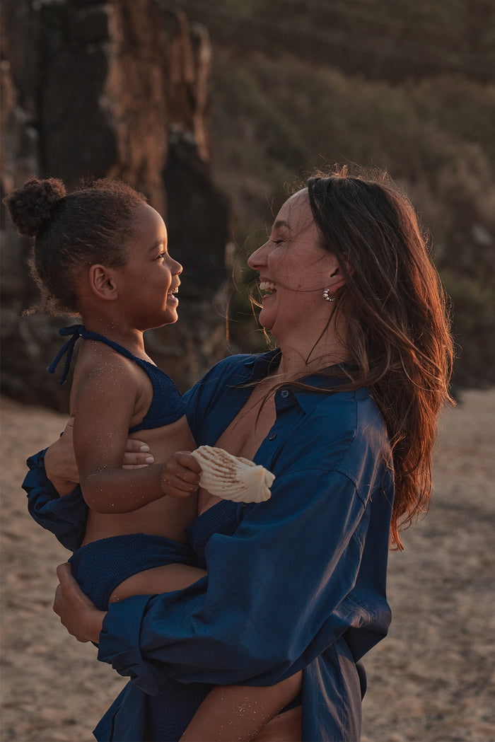 Mother laughing while holding her smiling young daughter on a sandy beach; child holds a seashell.