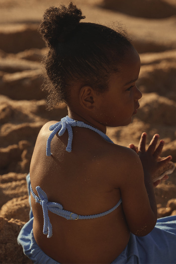 Back view of Palma top and Seychelles bottom set in Pool Crinkle on a child showing braided halter ties and gold ring detail on a sandy beach