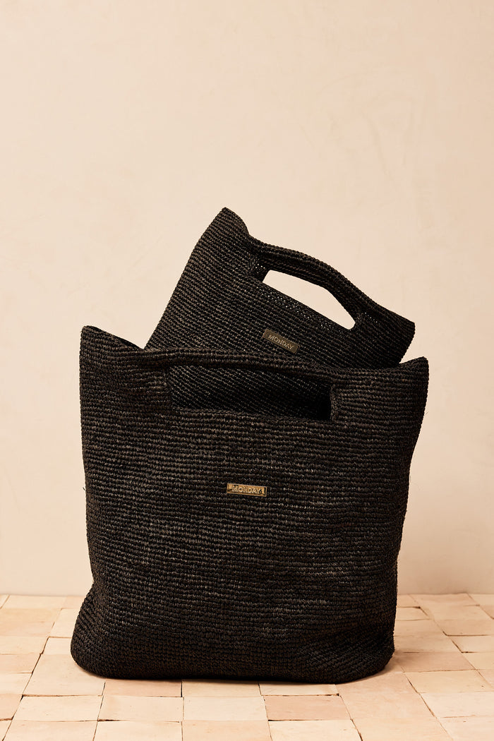 Front view of Paloma Bag Black Large showing woven raffia texture, cutout handles and small gold MONDAY logo on beige background