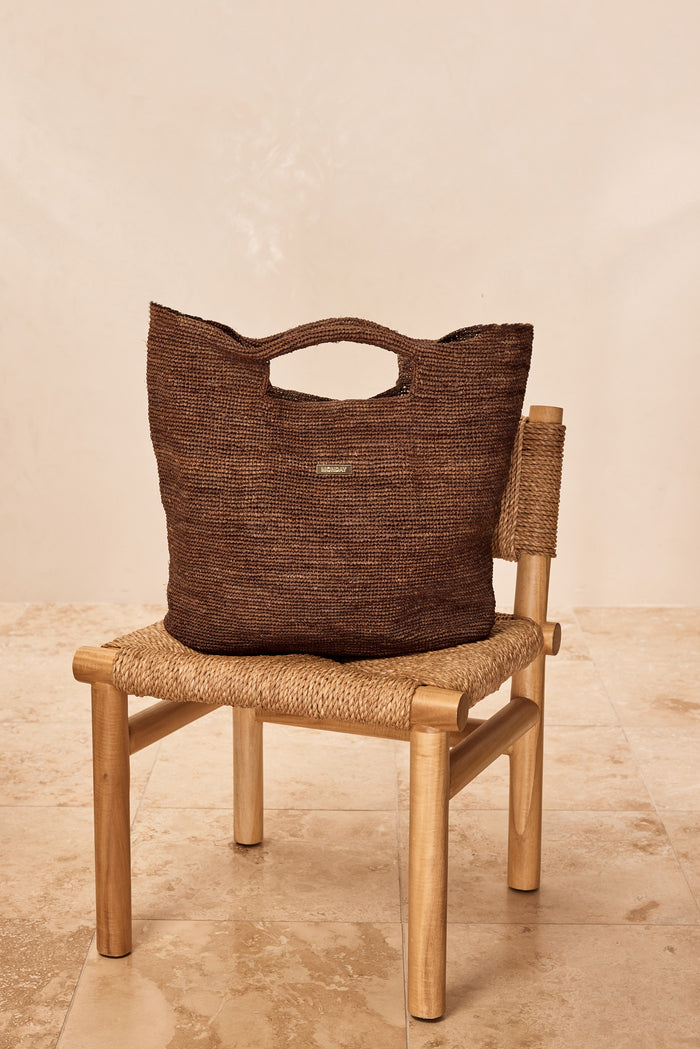Front view of Paloma Bag Large in espresso showcasing hand-crocheted raffia texture, cutout top handles, and gold MONDAY plaque on a woven chair