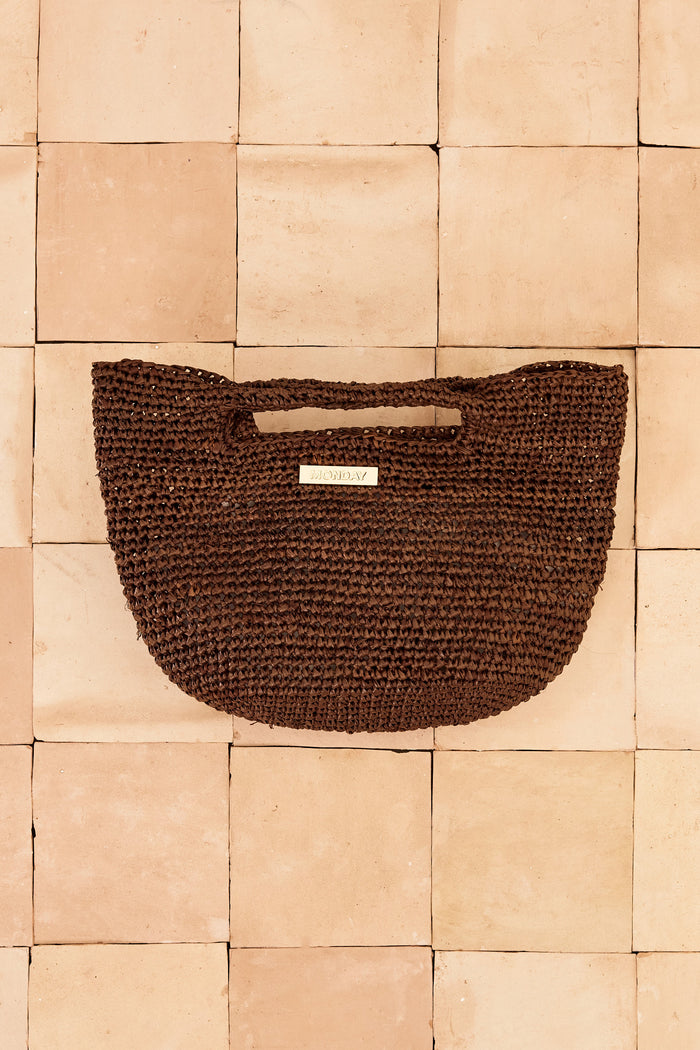 Top-down view of Mini Paloma bag showcasing hand-crocheted espresso raffia with a gold MONDAY logo plate on terracotta tiles