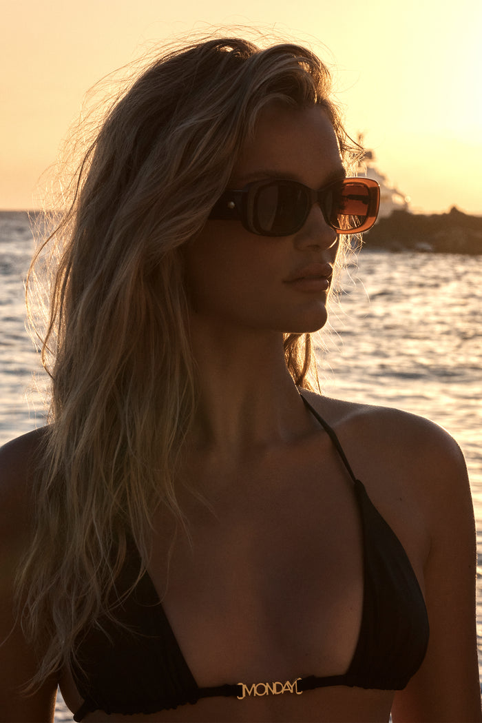 Three-quarter view of Piana sunglasses in brown showing square brown lenses and acetate frame against a seaside sunset background