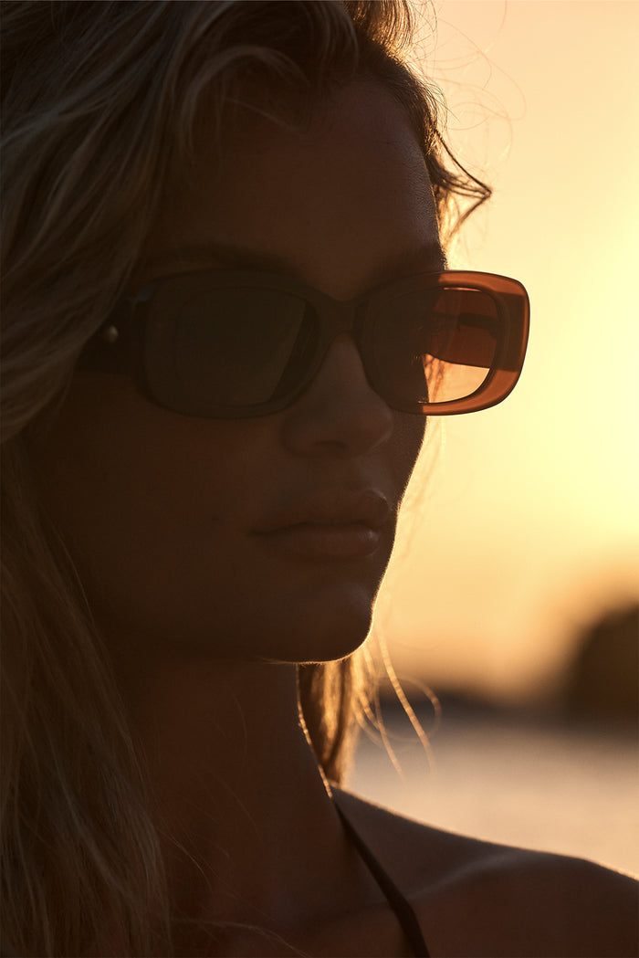 Model wearing Piana brown rectangular sunglasses at sunset, close-up showing warm backlight on acetate frame