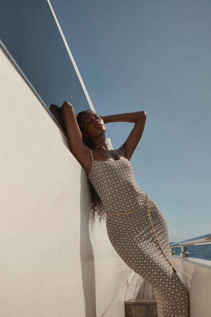 Model leaning back with arms raised wearing Provence tile-geo mesh slip dress in beige with gold chain belt on sunlit yacht