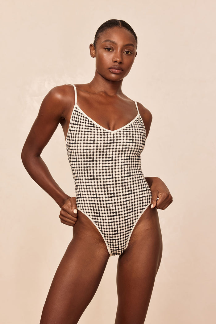 Front view of Ravello One Piece in black gingham showcasing scoop neckline, thin straps and high-cut leg on beige background