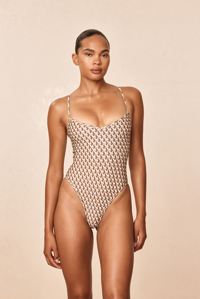 Front view of Ravello one-piece in Husk Geo showing scooped V neckline, thin adjustable straps and high-cut cheeky leg on beige background