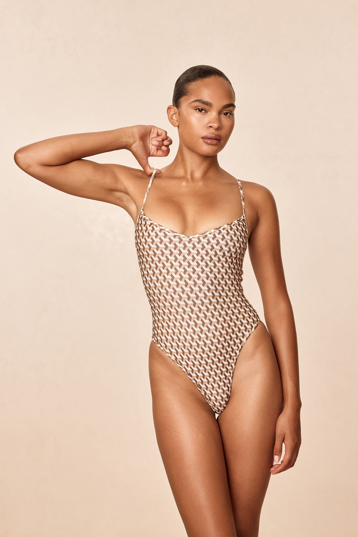 Front view of Ravello One Piece - Husk Geo showing wide V neckline, adjustable thin straps and high-cut leg on a neutral beige studio background