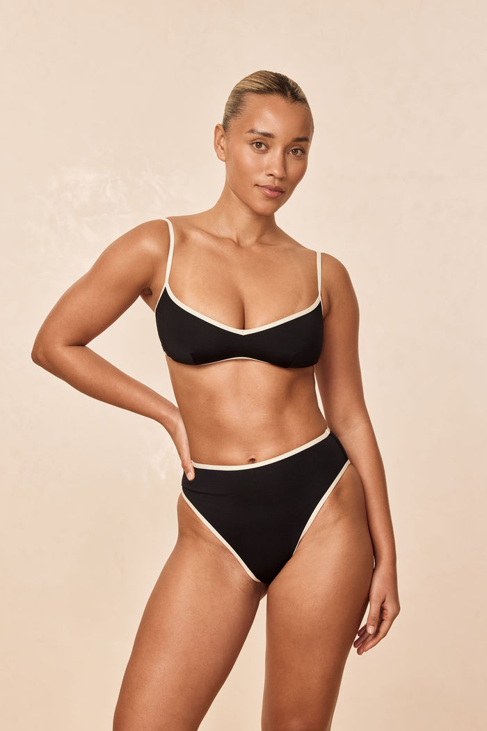 Front view of Ravello Top in black with ivory trim, thin straps and scooped neckline on model against beige background