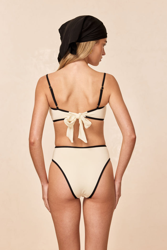 Back view of Ravello Top in ivory with black trim showcasing adjustable straps and tied bow back on a neutral beige background