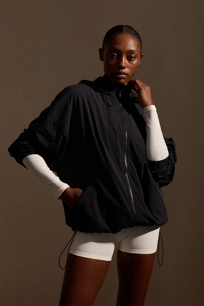 Three-quarter front of Rexford jacket in black on model showcasing hooded zip front, elastic cuffs and drawcord hem with neutral brown background