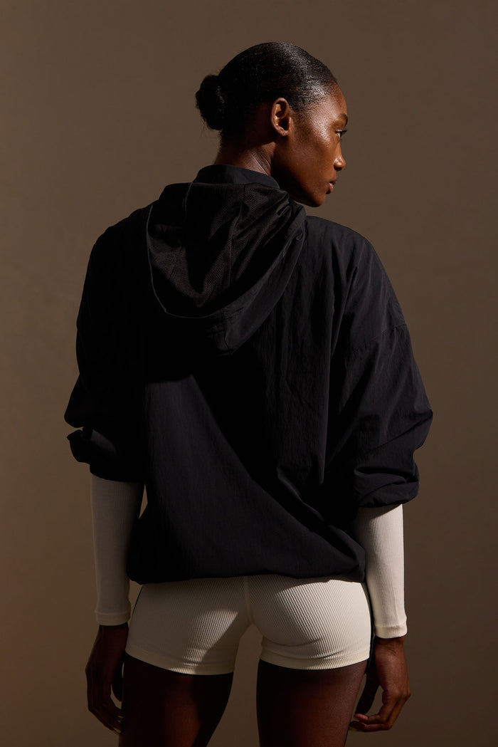 Back view of Rexford Jacket in black showcasing hood and oversized boyfriend fit with brown studio background