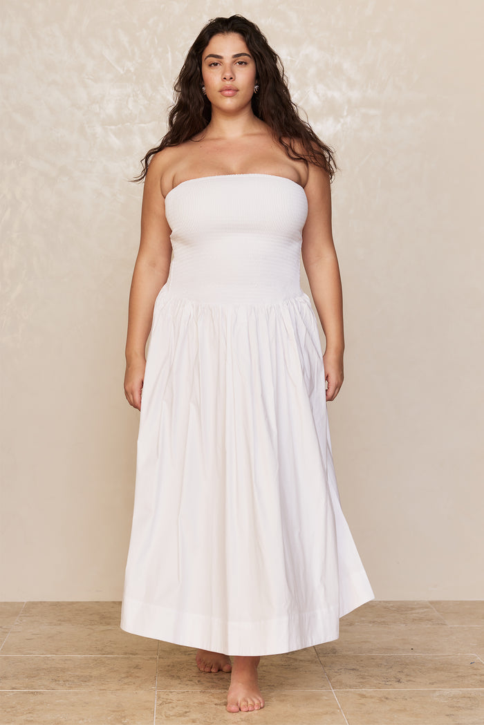 Model wearing white strapless smocked bodice and full ankle-length skirt, standing barefoot against a beige backdrop.
