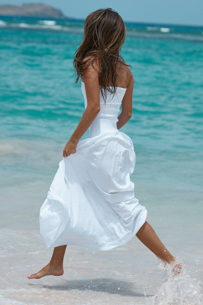 Back view of Rio dress in white showcasing smocked strapless bodice and gathered full maxi skirt with model at a turquoise ocean shoreline