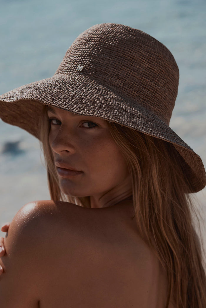 Rear three-quarter view of Rio Hat in Husk showing raffia weave and small gold M logo against beach background