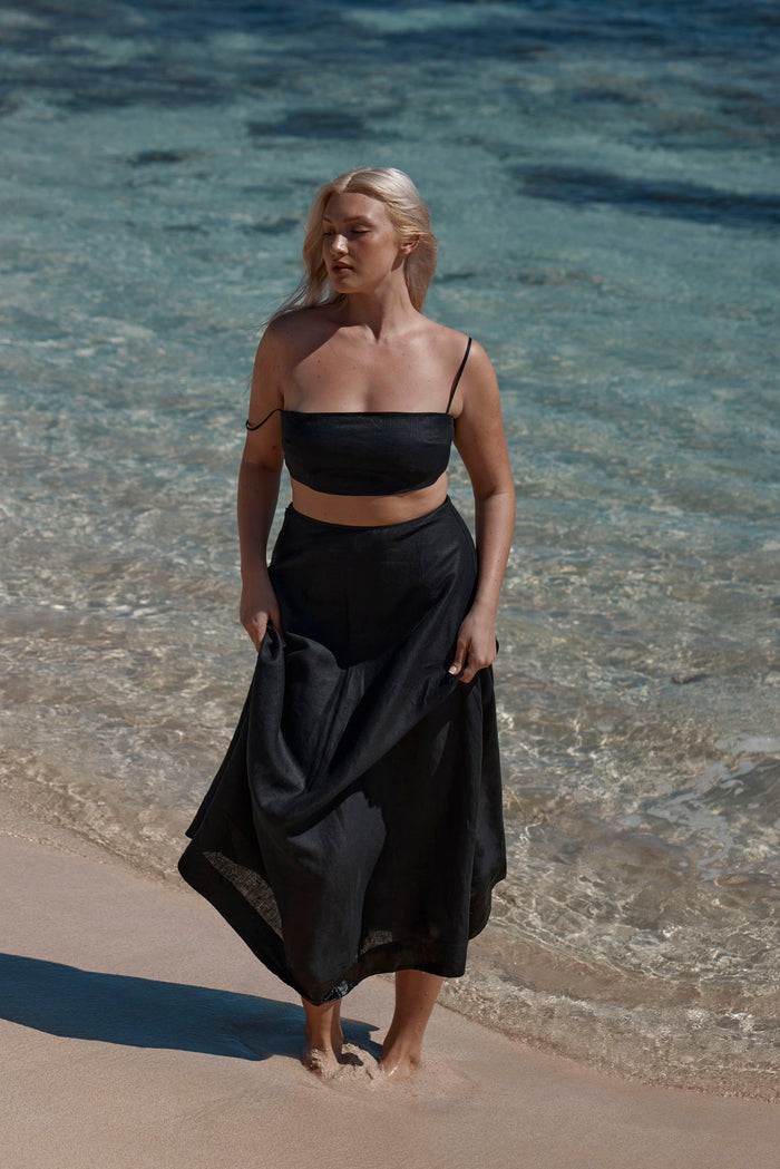 Front view of San Diego Top in black linen showcasing cropped silhouette and thin straps with model on sandy shore