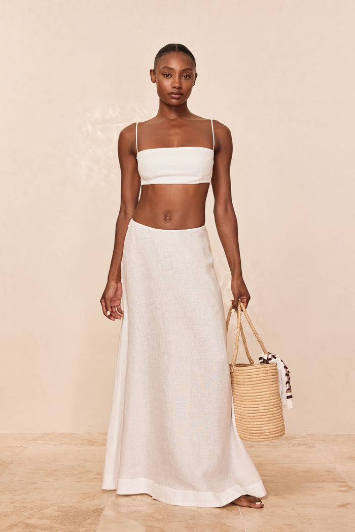 Model wearing white linen bandeau top and long A-line linen skirt, holding a woven straw bucket tote with knotted scarf.