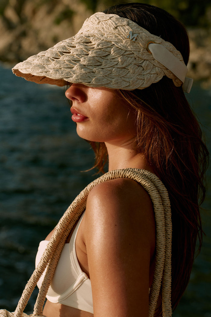 Woman in side profile at water's edge wearing woven straw visor with metal M emblem and carrying rope-handled bag.