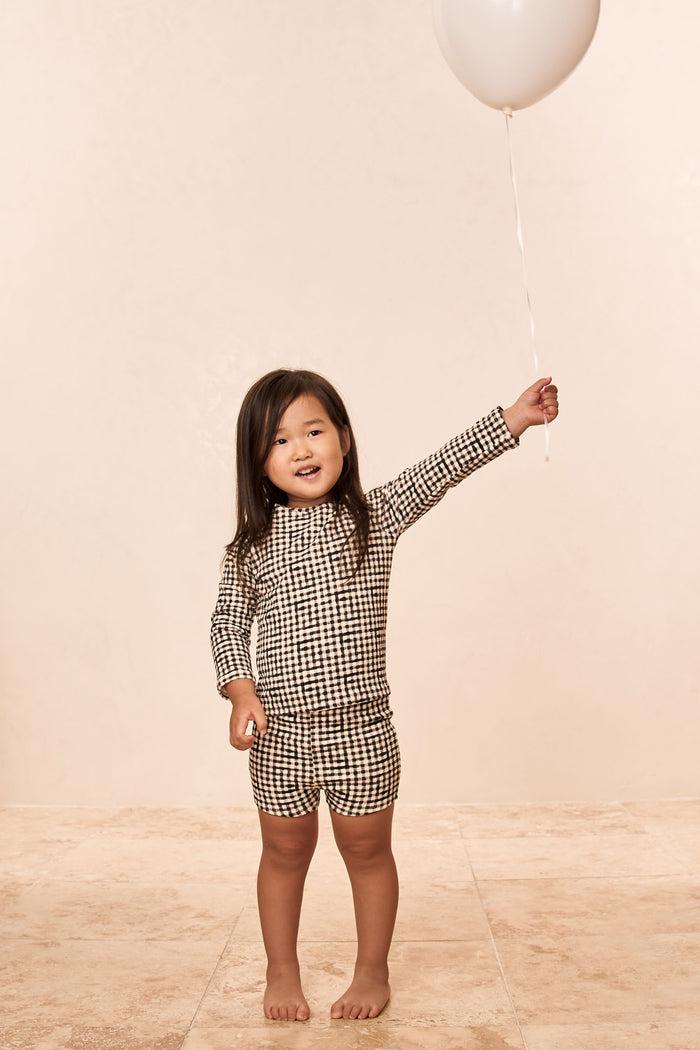 Front view of Santa Cruz Short Kids in Black Gingham on child model showing matching long-sleeve top and mid-thigh shorts on beige tile