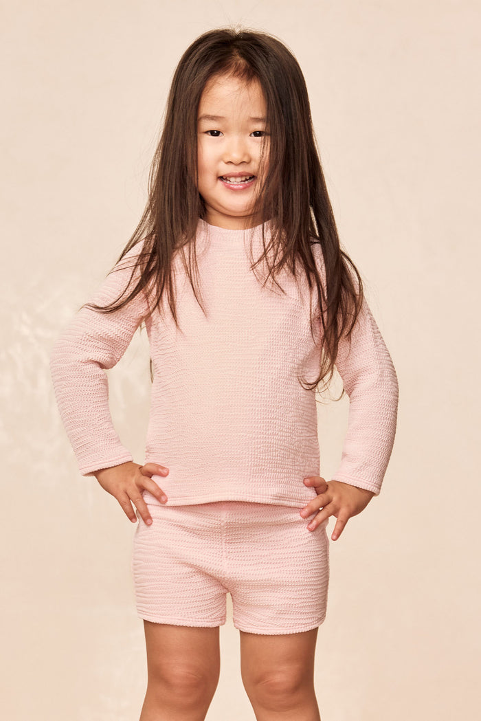 Front view of child wearing Santa Cruz kids shorts in powder pink crinkle showing textured fabric and elastic waist against beige background