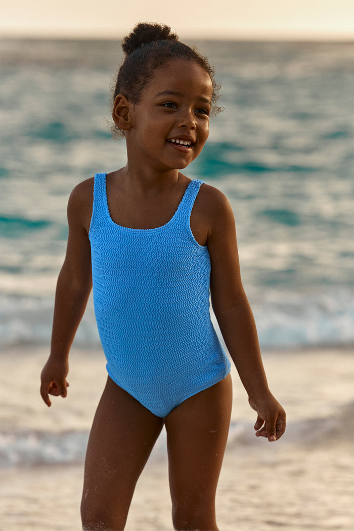 Three-quarter view of Sardinia one-piece in Azure Crinkle showcasing textured crinkle fabric and scoop neckline with ocean waves and sandy beach background