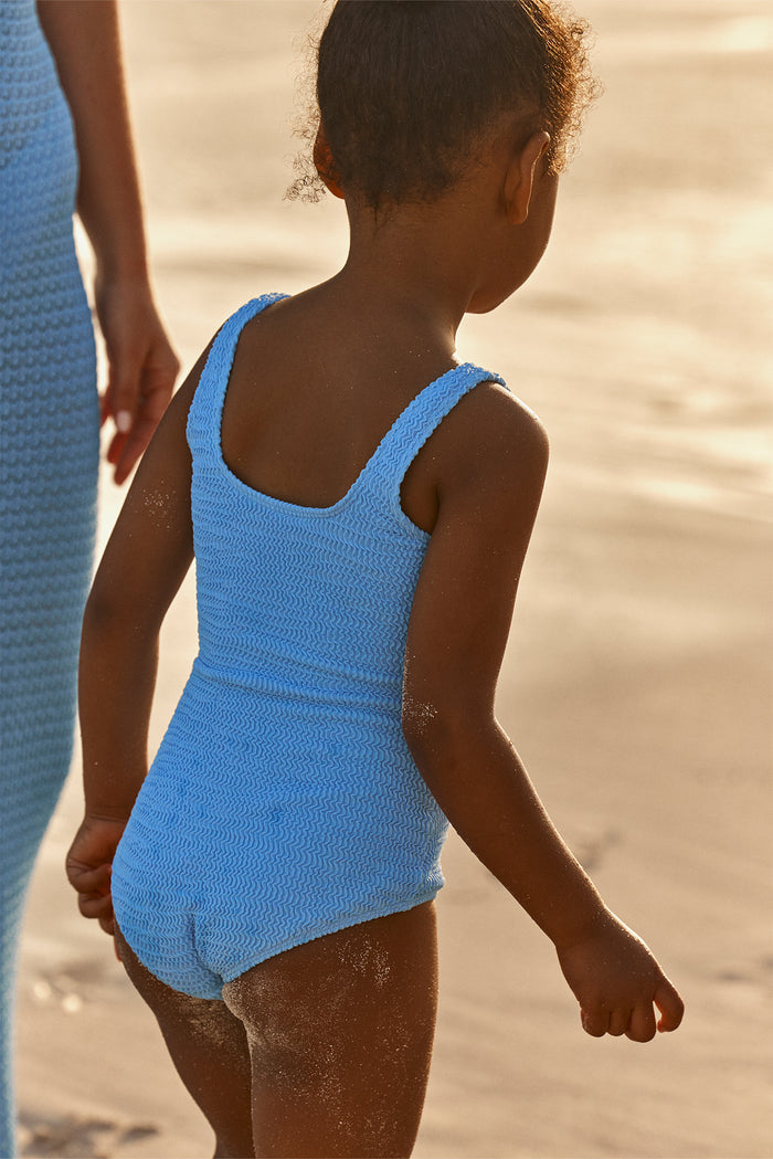 Rear view of Sardinia one-piece in Azure Crinkle showcasing crinkle texture and scoop back with wide straps on a sandy beach