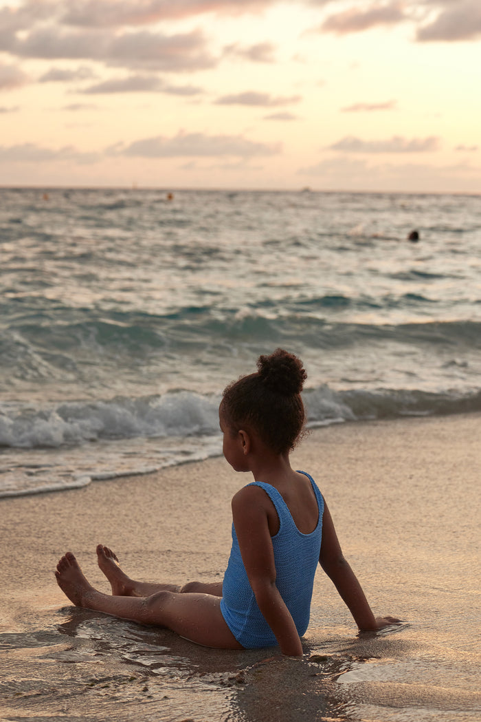 Three-quarter rear view of Sardinia one-piece in Azure Crinkle showing textured crinkle fabric and wide straps on a child at the beach