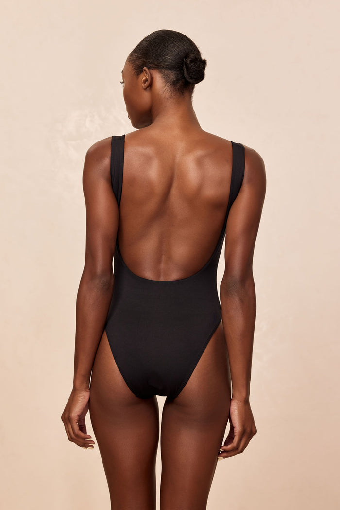 Back view of Sardinia one-piece swimsuit in black showing deep scoop back and high-cut leg on model against beige background