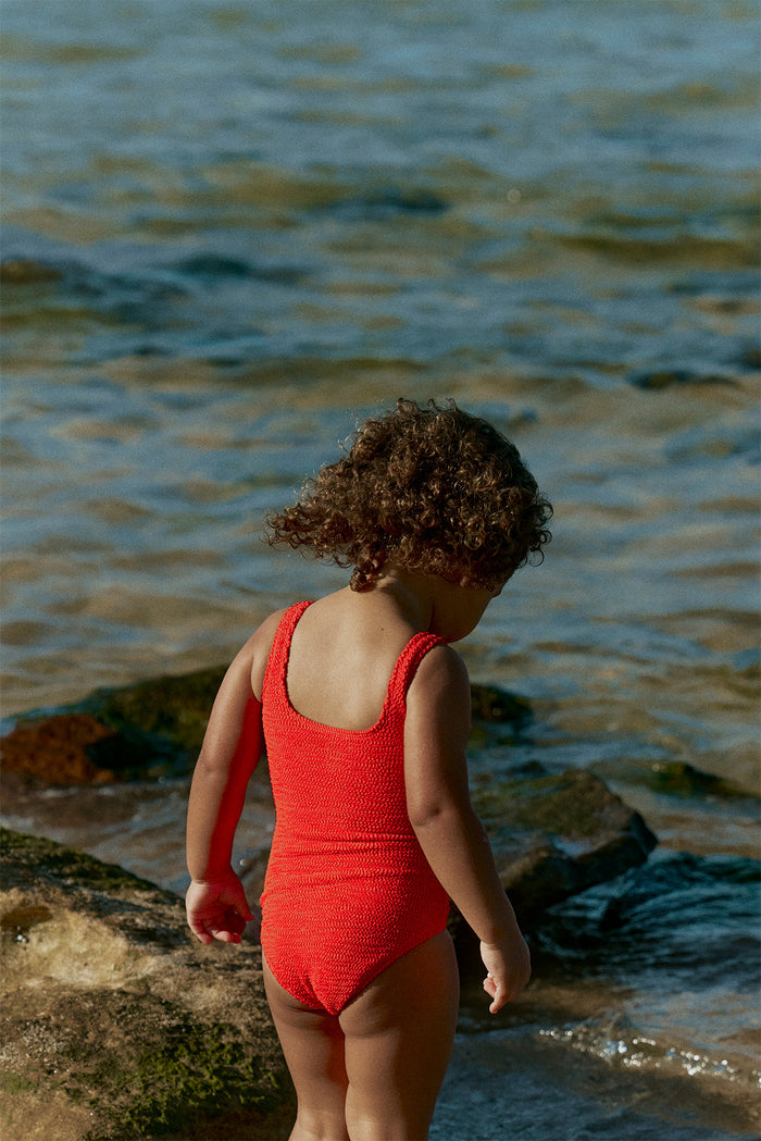 Back view of Sardinia One Piece in Chili Pepper Crinkle showcasing textured crinkle knit and scoop-back on a child at a rocky shoreline