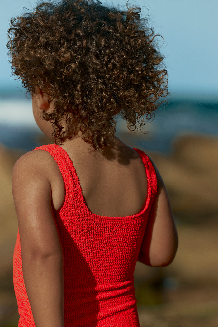 Child with curly hair seen from behind wearing a red textured sleeveless swimsuit at the beach
