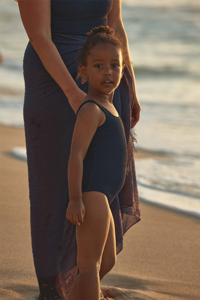 Side view of Sardinia One Piece in Midnight Crinkle on a child model showcasing crinkle texture with sunset beach background