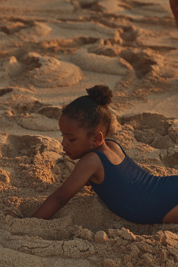 Side view of Sardinia One Piece in Midnight Crinkle showcasing crinkle texture and scoop neckline with sandy beach at sunset