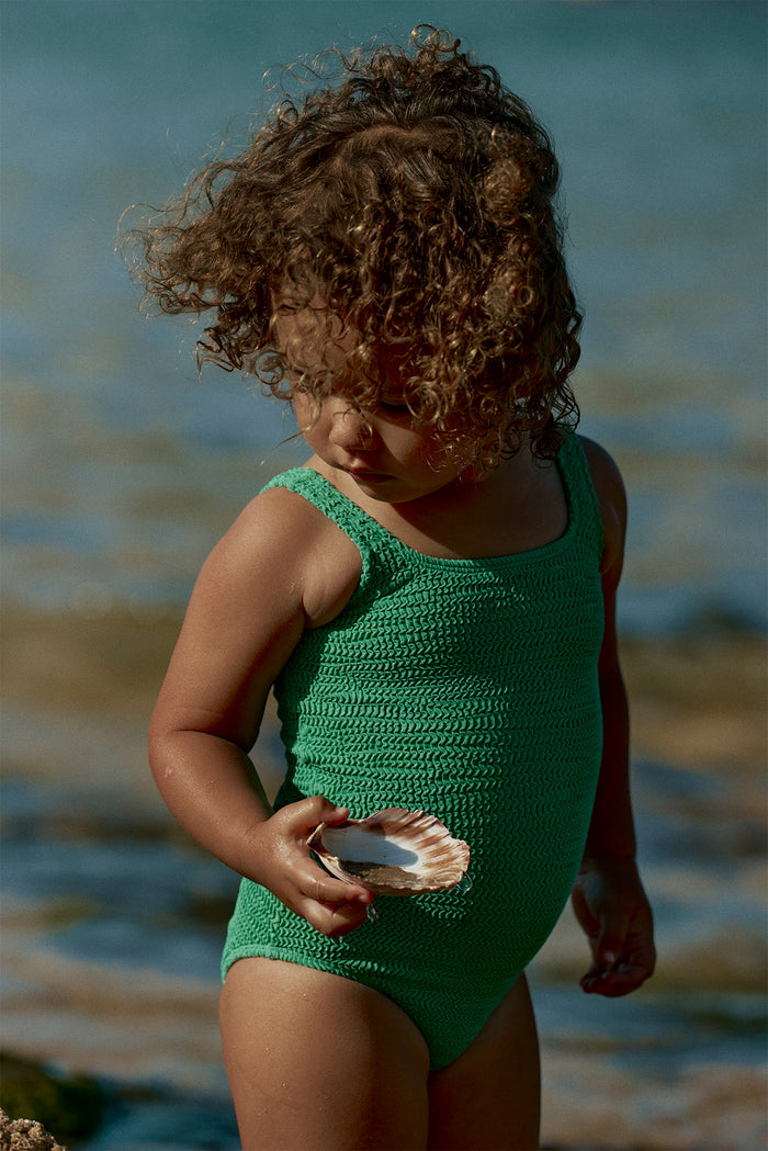 Three-quarter view of Sardinia One Piece in Pond Crinkle showing textured crinkle knit and scoop neckline on a toddler at the shoreline