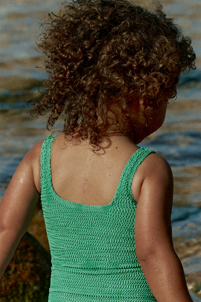 Back view of Sardinia One Piece in Pond Crinkle showcasing crinkle knit texture and full back coverage on a child at the shoreline