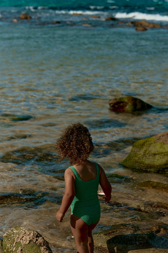 Back view of Sardinia one-piece swimsuit in Pond Crinkle showing crinkle texture and coverage worn by child on rocky shore